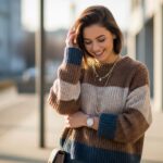 Mladá žena v teplom svetríku stojaca vonku v mestskom prostredí so zrelaxovaným úsmevom.