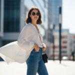 Mladá žena v voľnej blúzke a džínsach pózuje na mestskom pozadí za denného svetla.